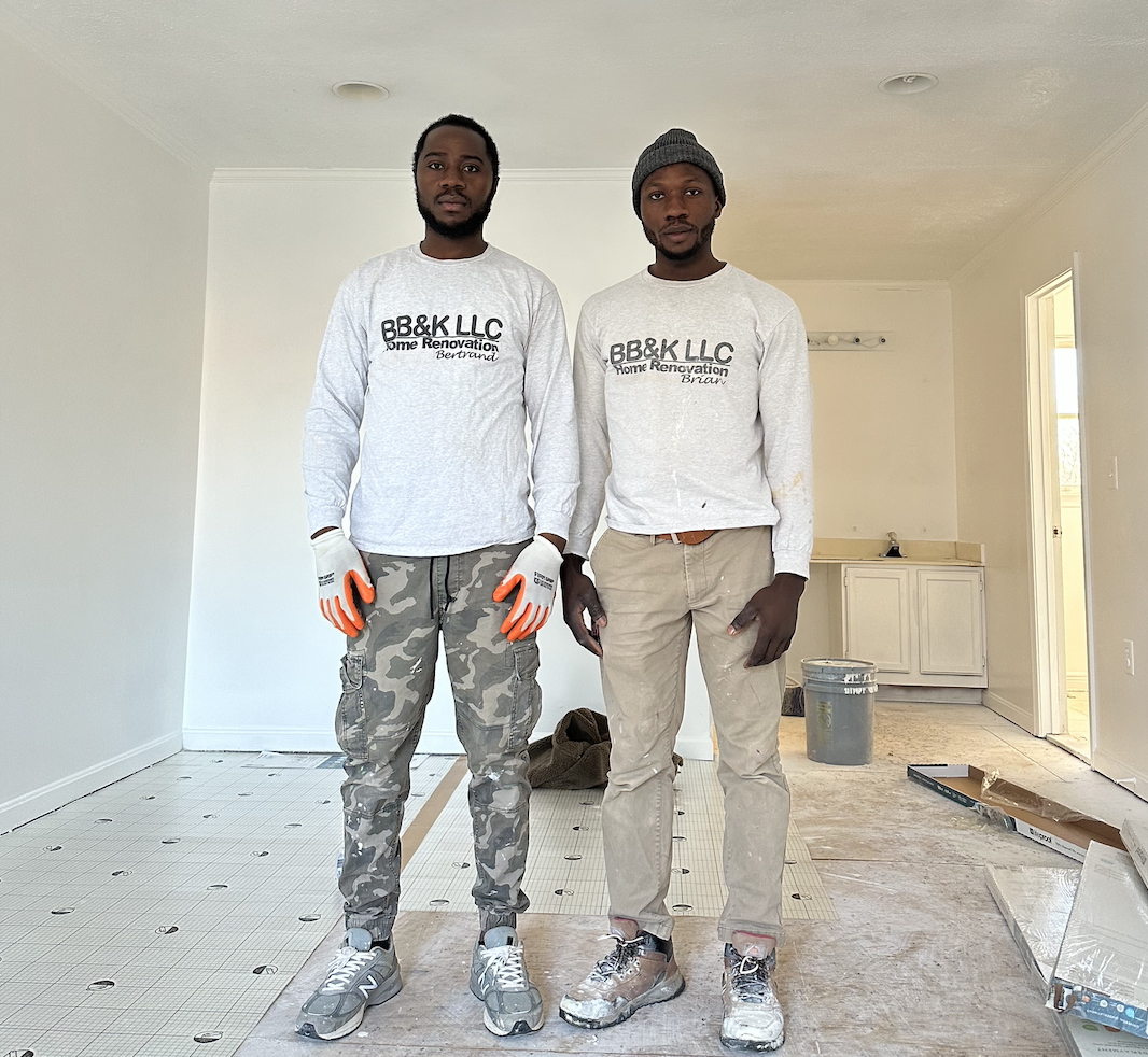 Two workers in white shirts and work pants standing in a renovation room with tools and dust on the floor
