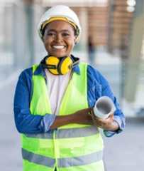 Construction worker in hard hat and safety vest holding a pipe, smiling in a workshop.
