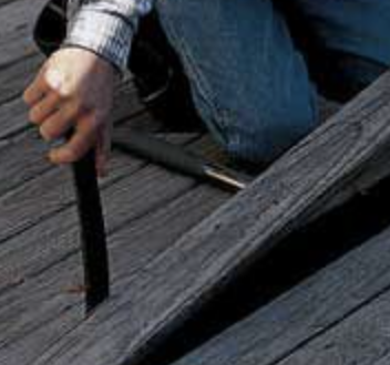 Hand gripping a black tool near a wooden deck gap, with a metal bar beside it