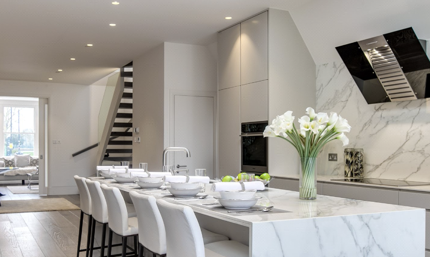 Modern white kitchen with marble island, dining chairs, staircase, and vase of white flowers