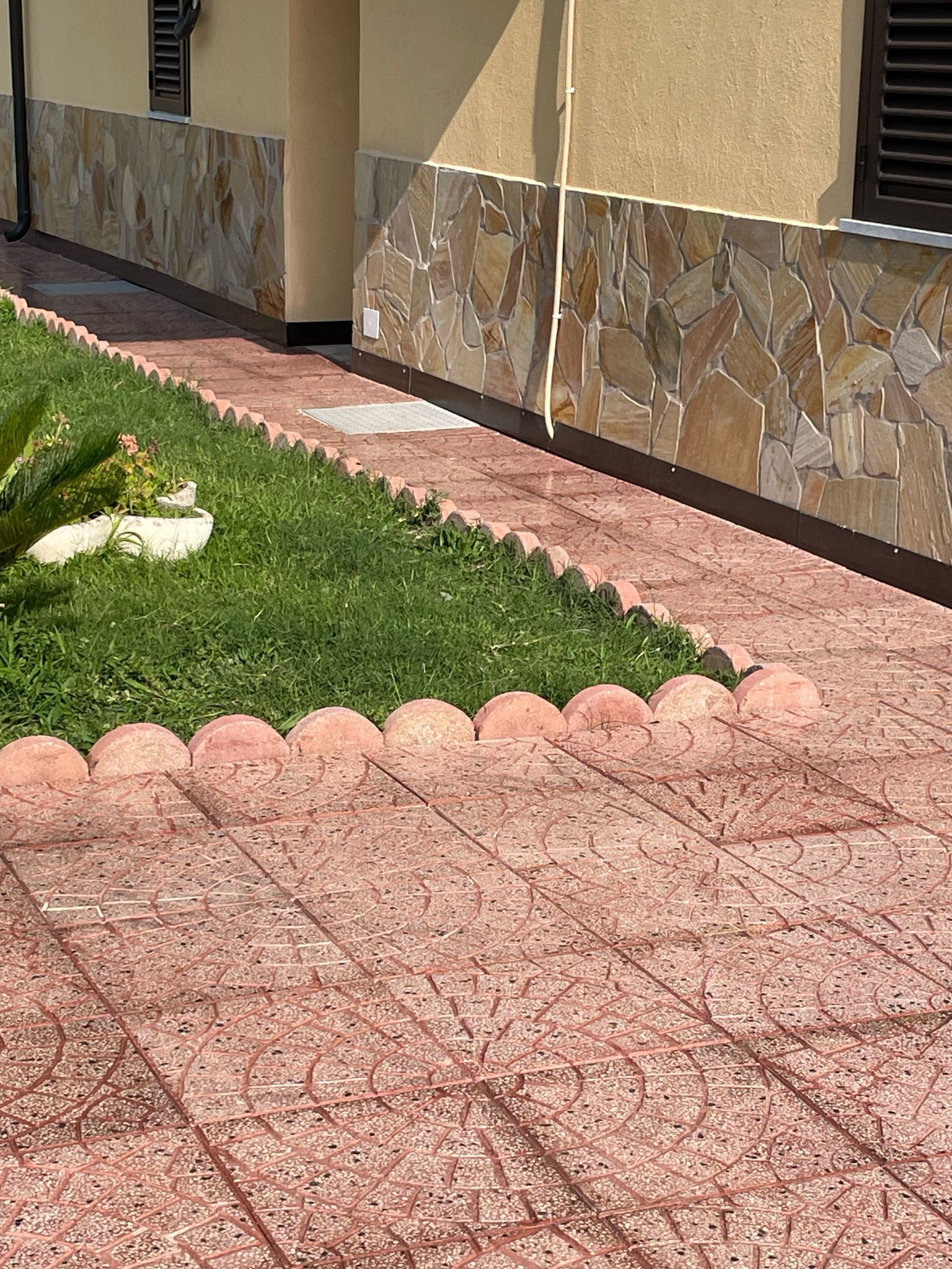 Un sentiero lastricato con bordature decorative costeggia un prato verde. Sullo sfondo si trova un muro con dettagli in pietra.