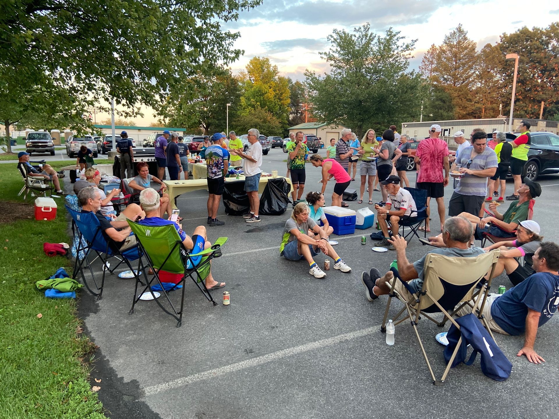 Group of people in a parking lot, some in chairs, socializing after an event. Trees and parked cars in the background.