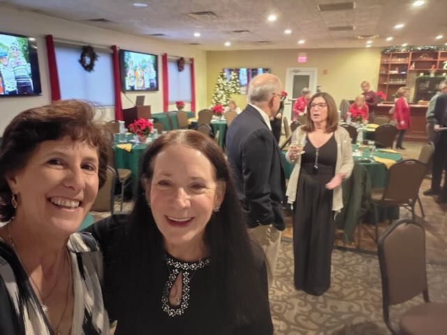 Two women smiling for a selfie at a holiday party; others socialize at tables, decorated with green and red accents.