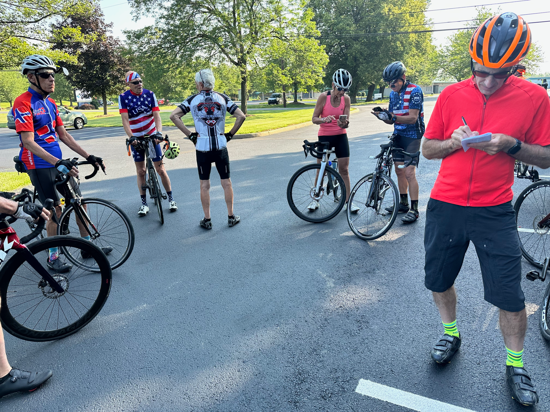 Cyclists in cycling gear gather outdoors, some looking at a person with a clipboard; park setting.