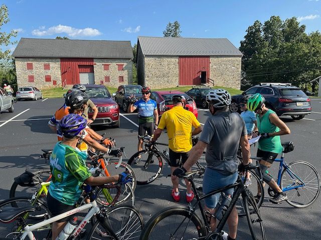 Cyclists gather in a parking lot, near two stone barns with red doors, preparing for a ride.