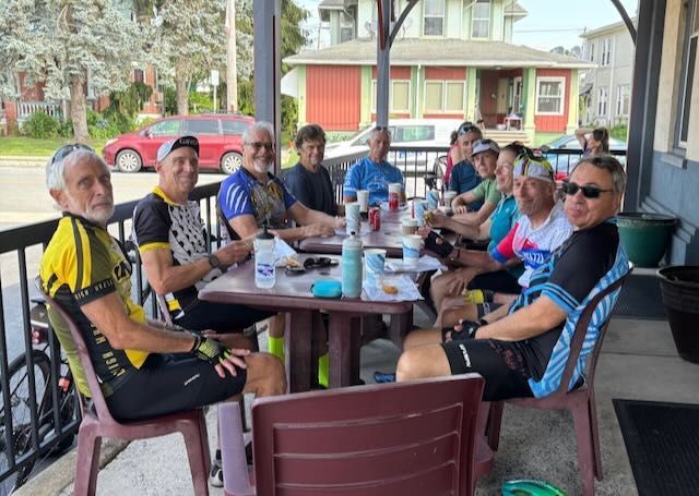 Cyclists in colorful jerseys sit at a table on a porch, eating and drinking.