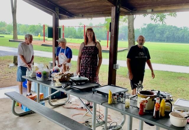 Four people at a picnic table under a shelter, cooking outdoors with a park background.