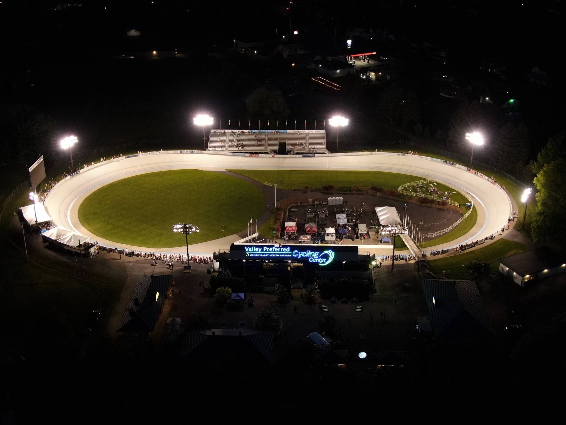 Nighttime aerial view of a brightly lit racetrack, with bleachers and a stage, set against a dark landscape.