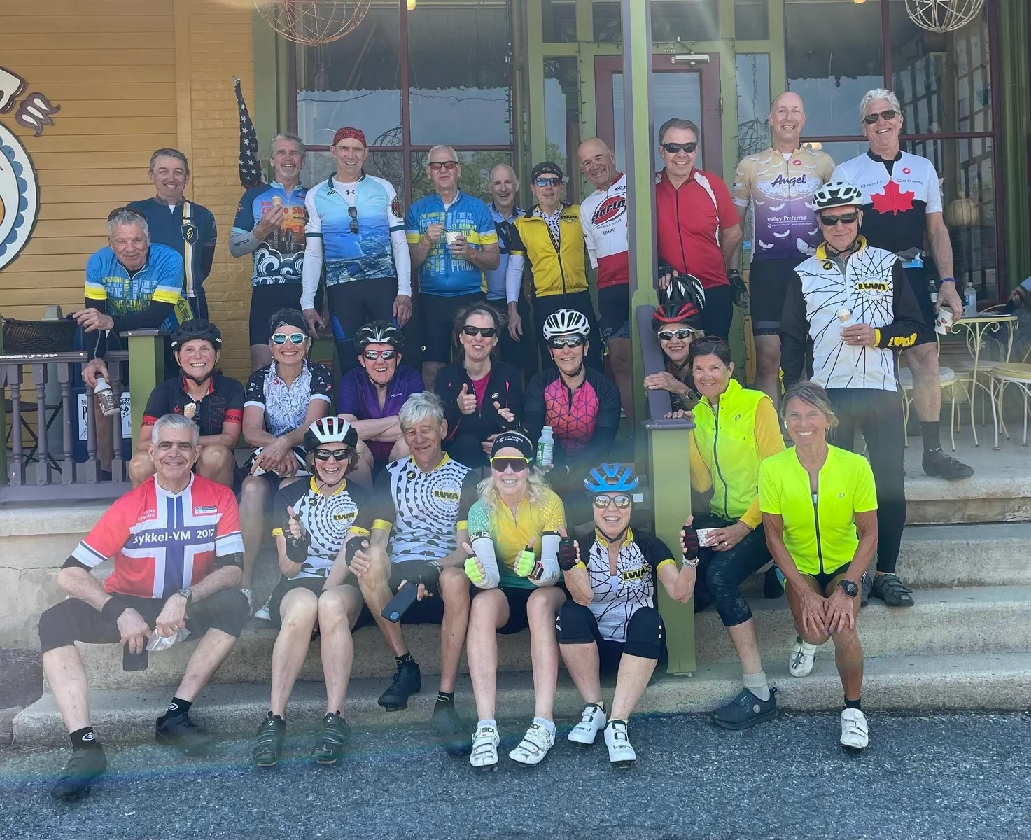 Group of cyclists pose on steps outside a building. Many wear cycling jerseys, smiling. Sunny day.
