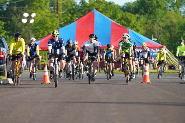 Cyclists racing on a paved road toward a red and blue tent; orange cones mark the course.