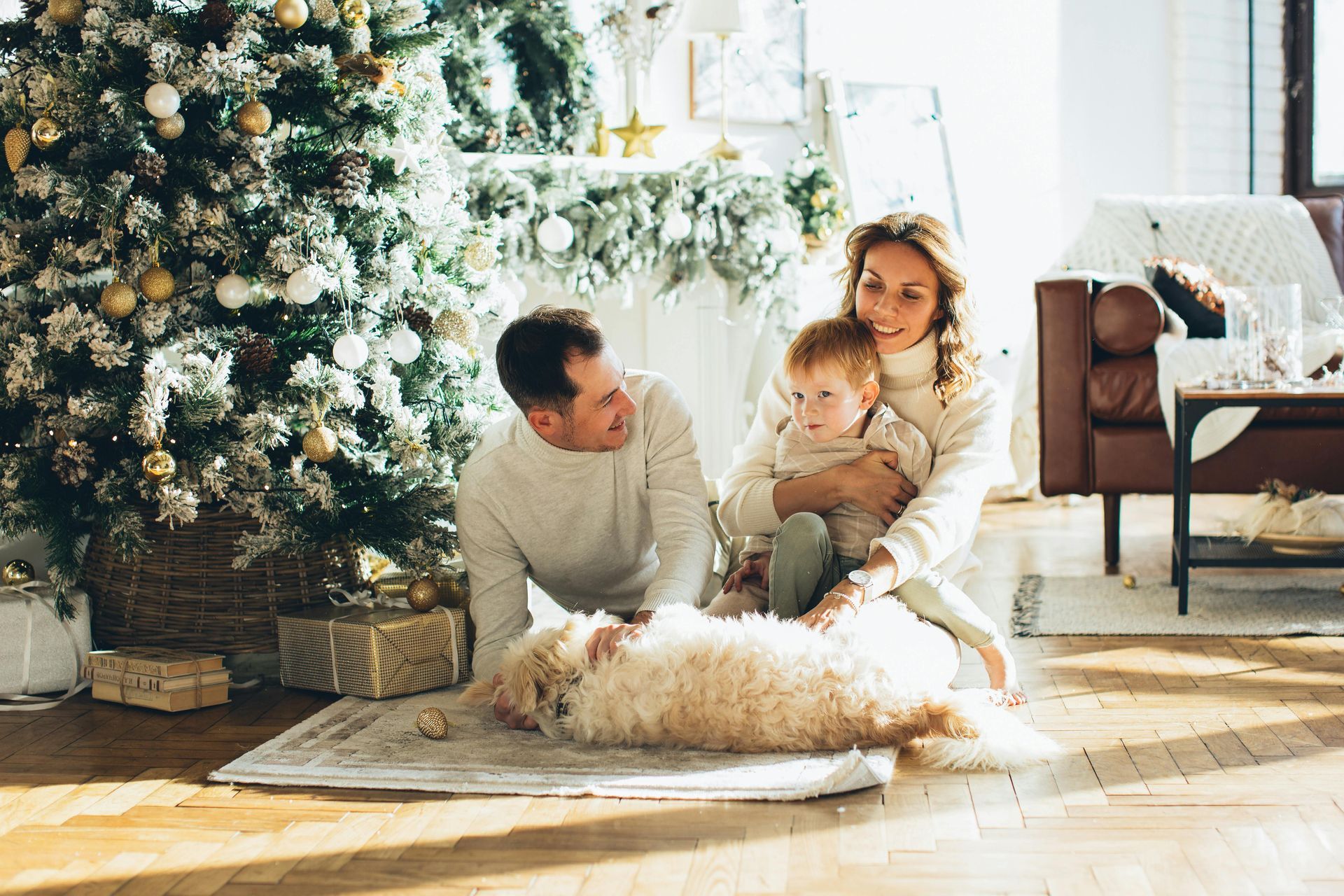 Family with a toddler and dog in front of a Christmas tree, smiling, in a cozy room.
