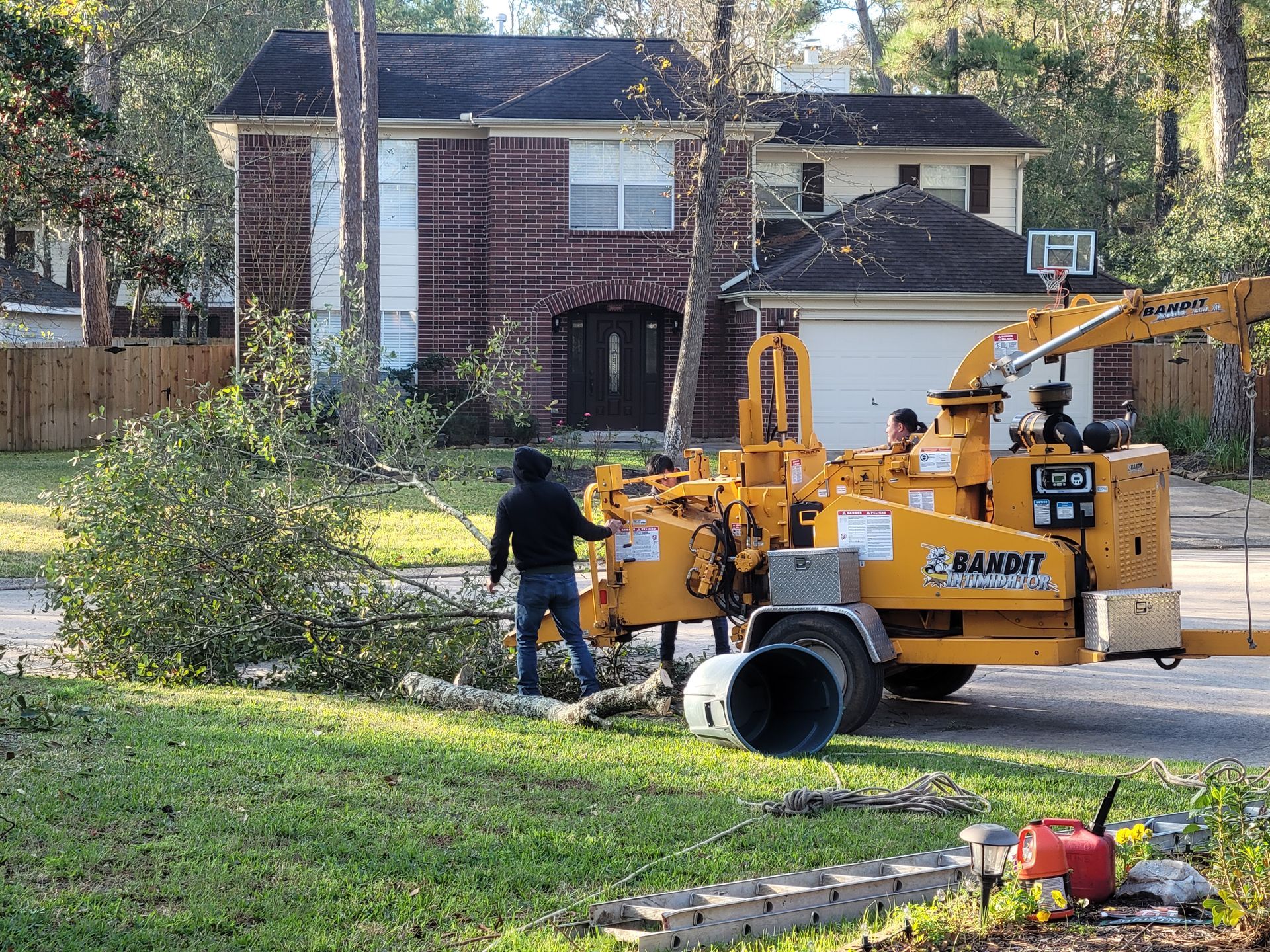 Yellow utility truck and worker trimming trees in front of a brick house on a suburban street.