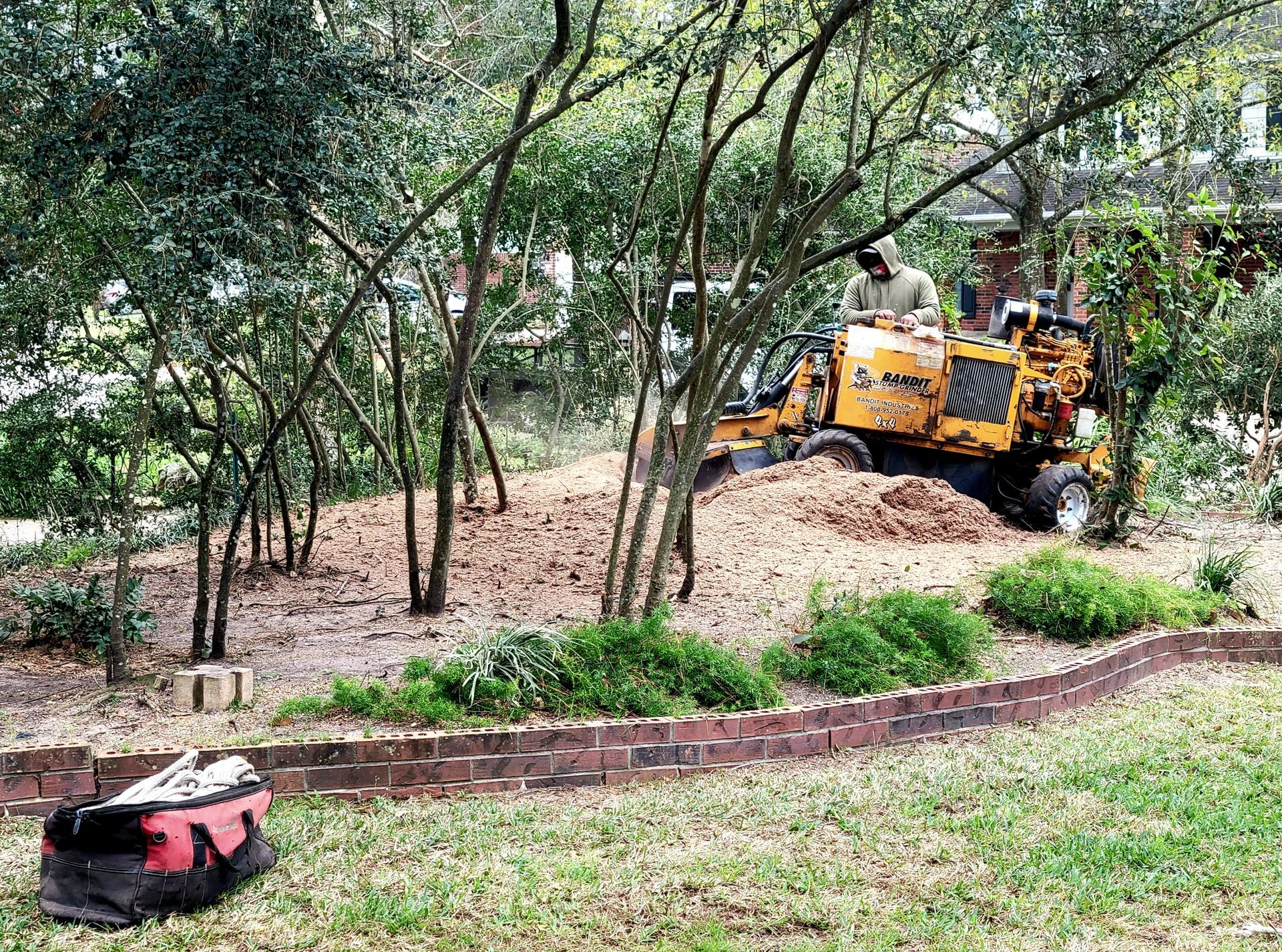 Yellow utility vehicle among trees in a wooded yard, with mulch, shrubs, and a red mower in the foreground