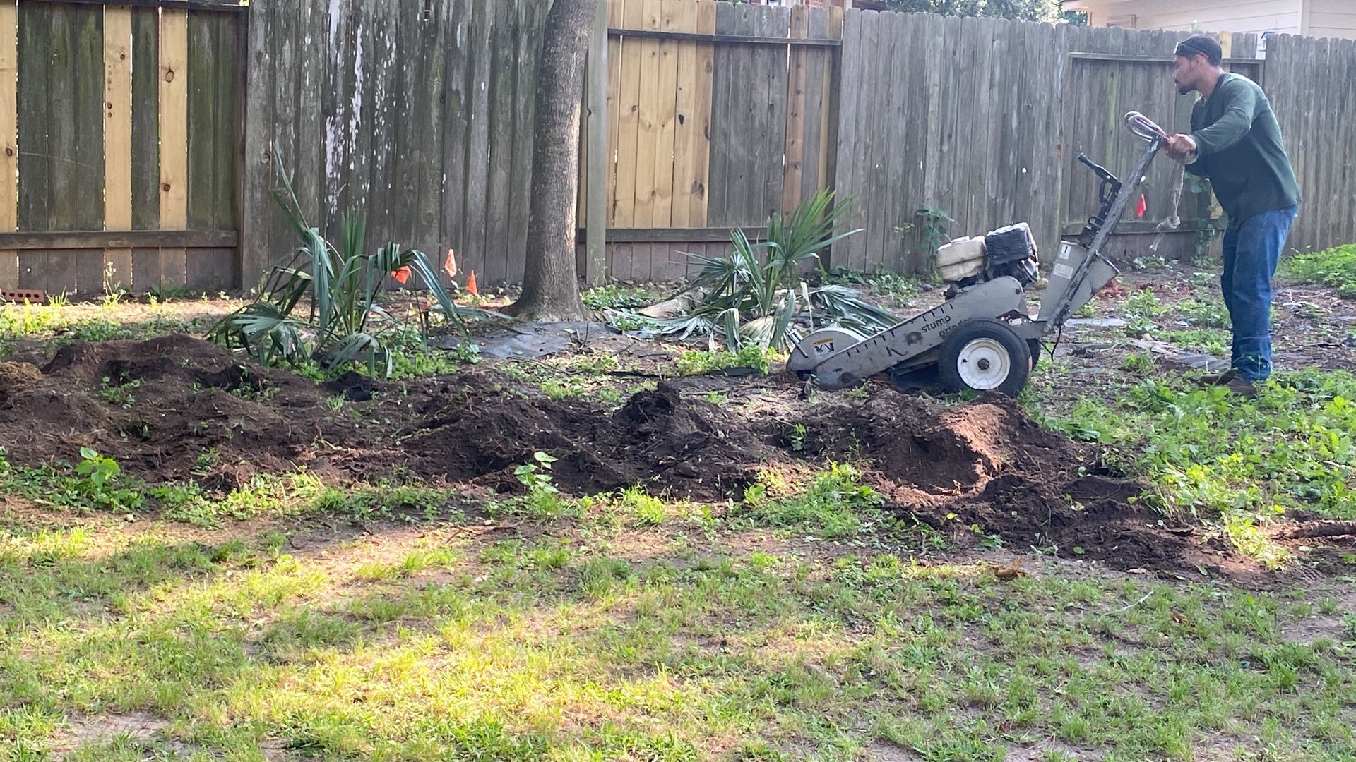 Person operating a tiller in a backyard, turning over dark soil near a wooden fence.