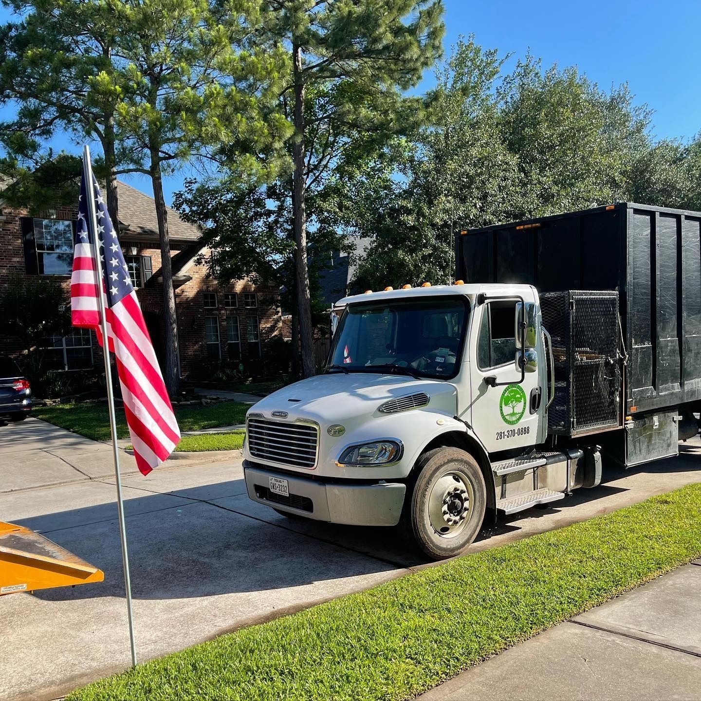 White utility truck parked by a sidewalk with an American flag and black trailer under trees.
