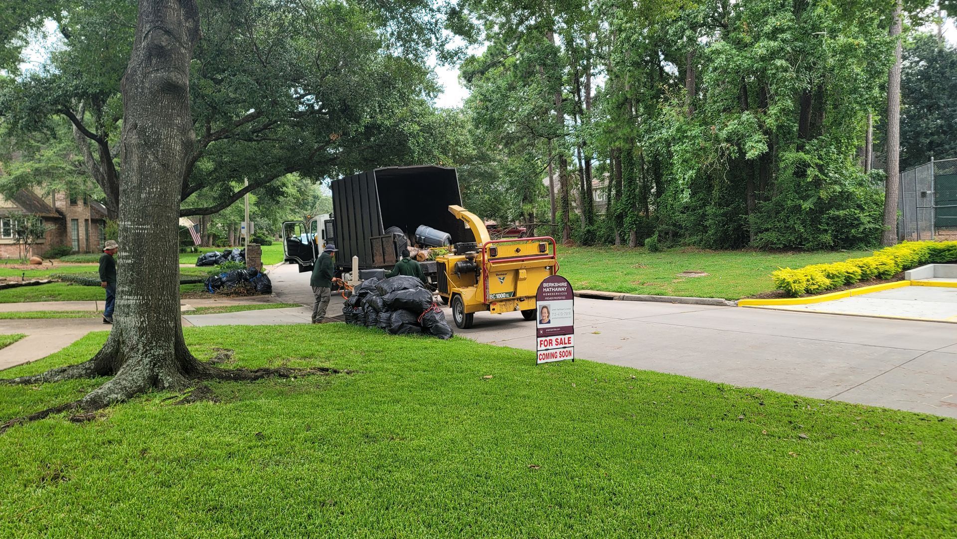 Yellow roadwork machine on a residential street beside a lawn and trees, with workers nearby.