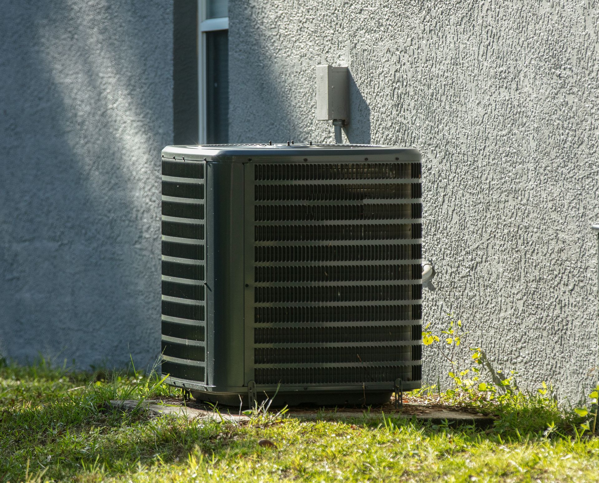 Outdoor air conditioner unit beside a gray wall on a sunny lawn Outdoor air conditioner unit beside a gray wall on a sunny lawn