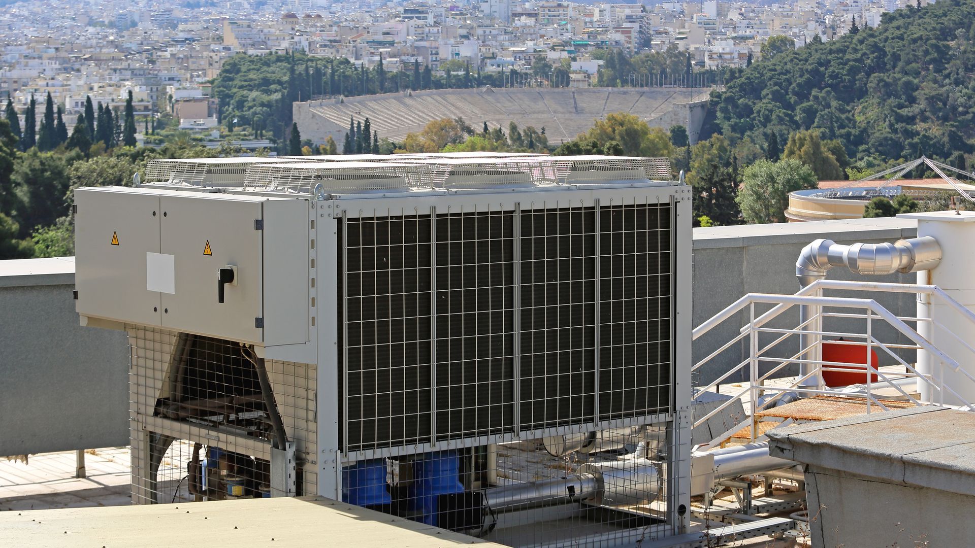 Large rooftop HVAC unit on a building, with cityscape and trees in the background. Large rooftop HVAC unit on a building, with cityscape and trees in the background.