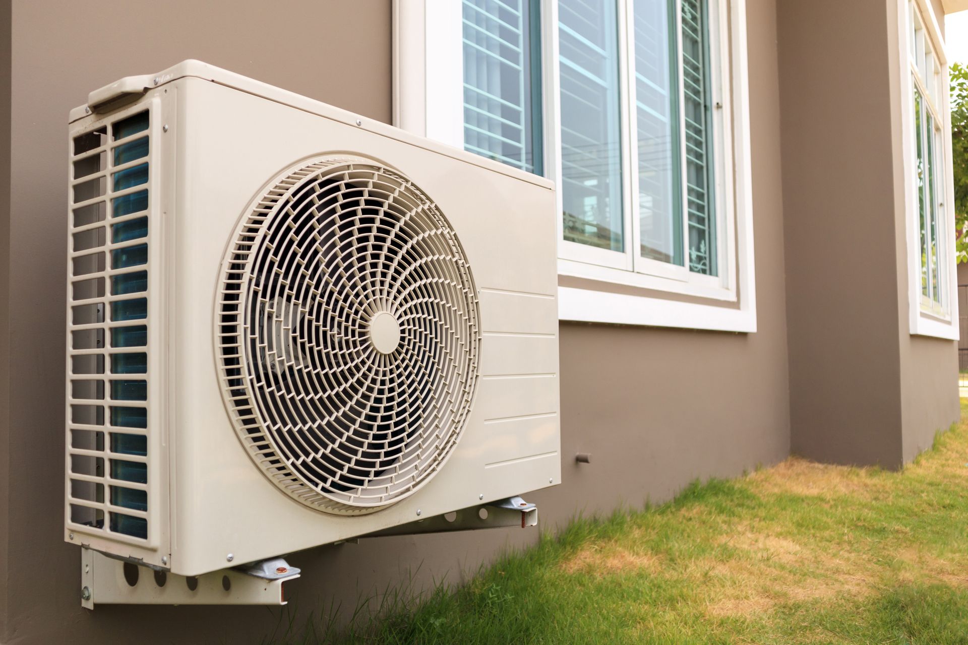 Outdoor AC unit mounted beside a house with windows and a grassy yard Outdoor AC unit mounted beside a house with windows and a grassy yard