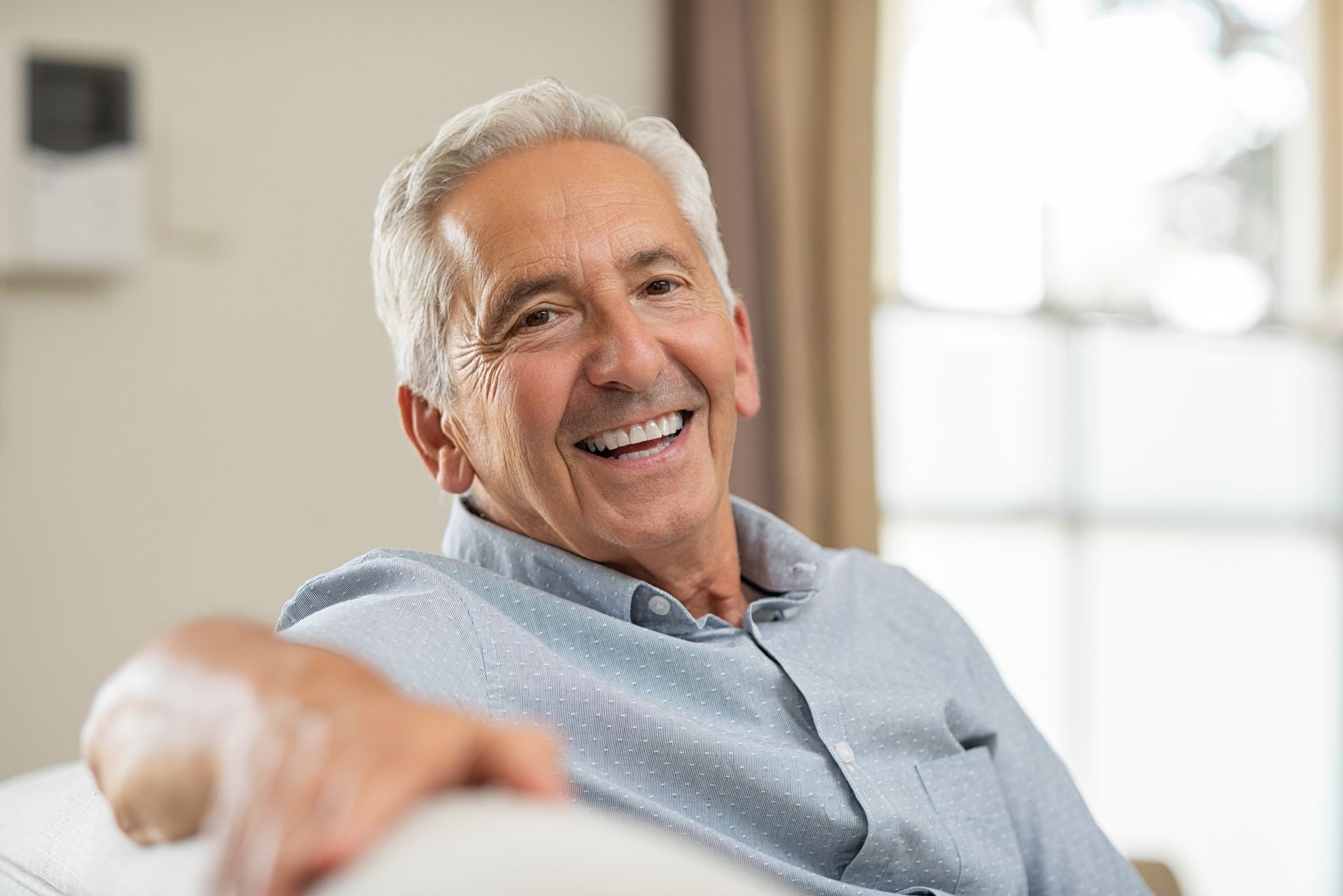 Smiling senior man in a light blue shirt sits on a couch indoors, looking at the viewer.