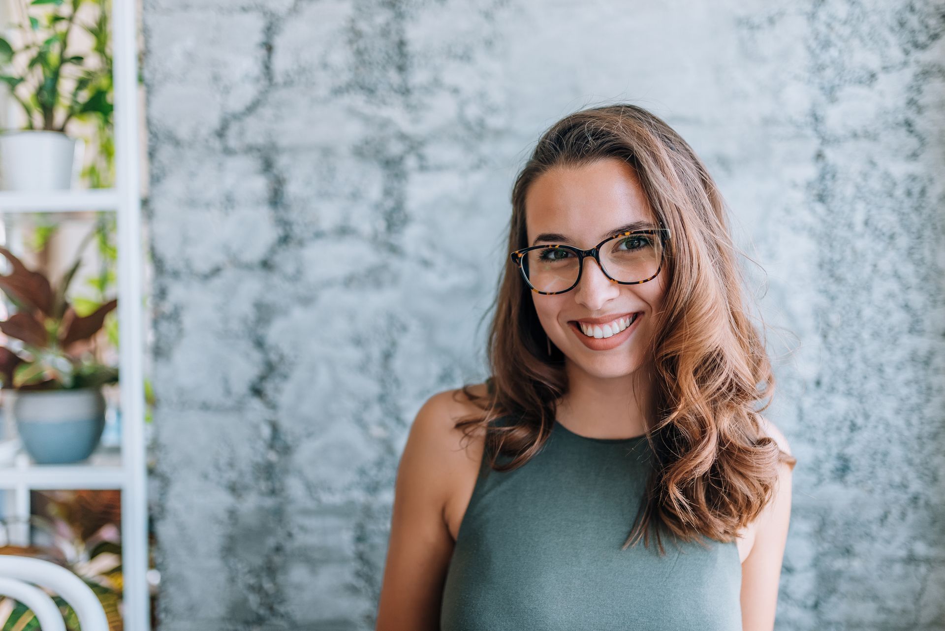 Woman with glasses smiling, standing in front of a gray brick wall and plants.