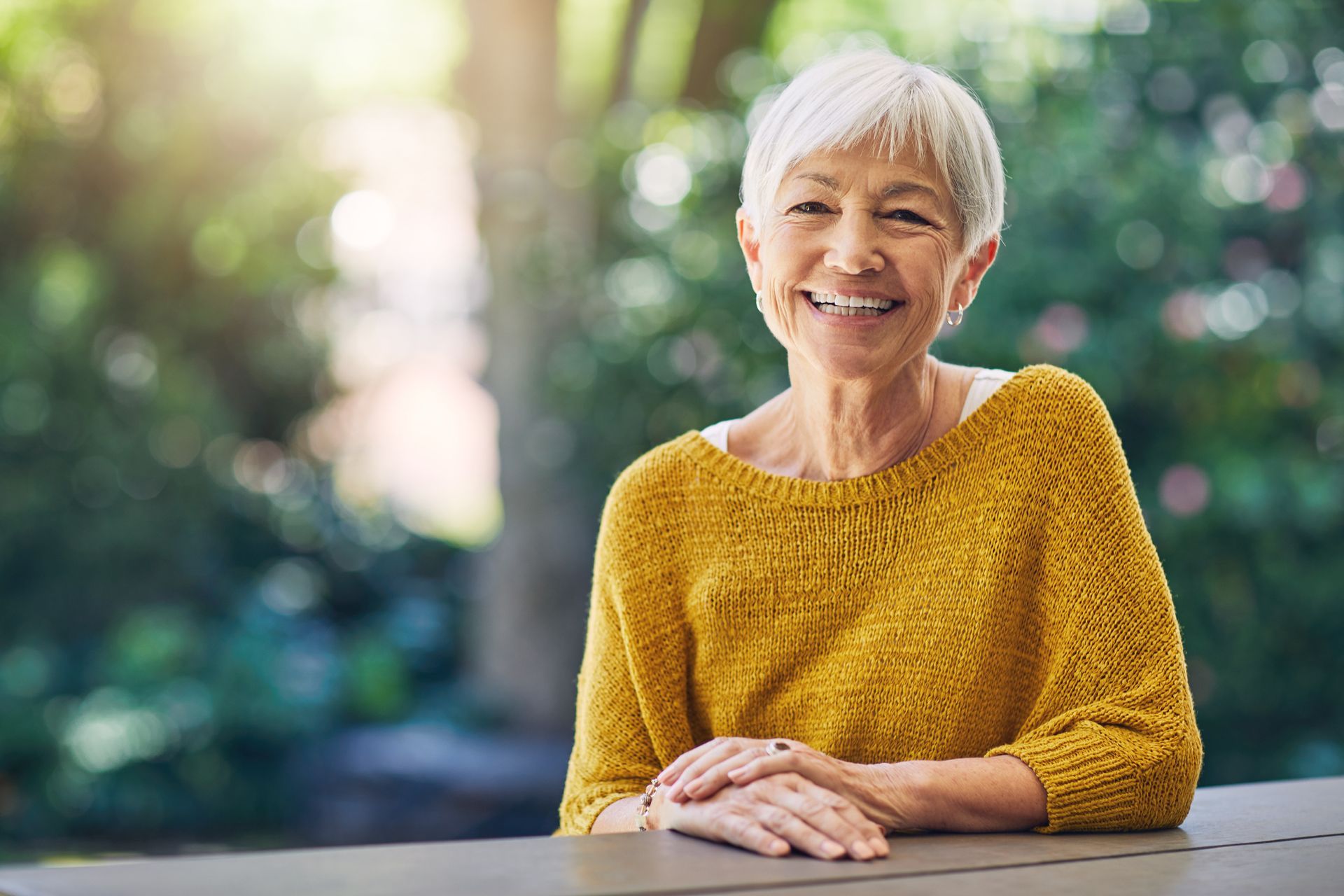 Smiling older woman with short gray hair in a yellow sweater, sitting outside, smiling at the camera.