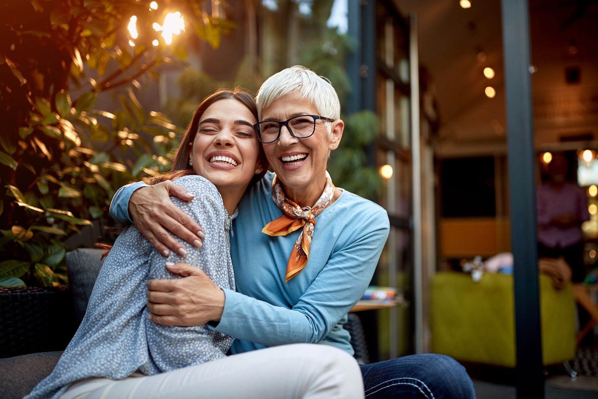 Smiling woman hugs younger woman outdoors; warm sunlight, plants, cafe setting.