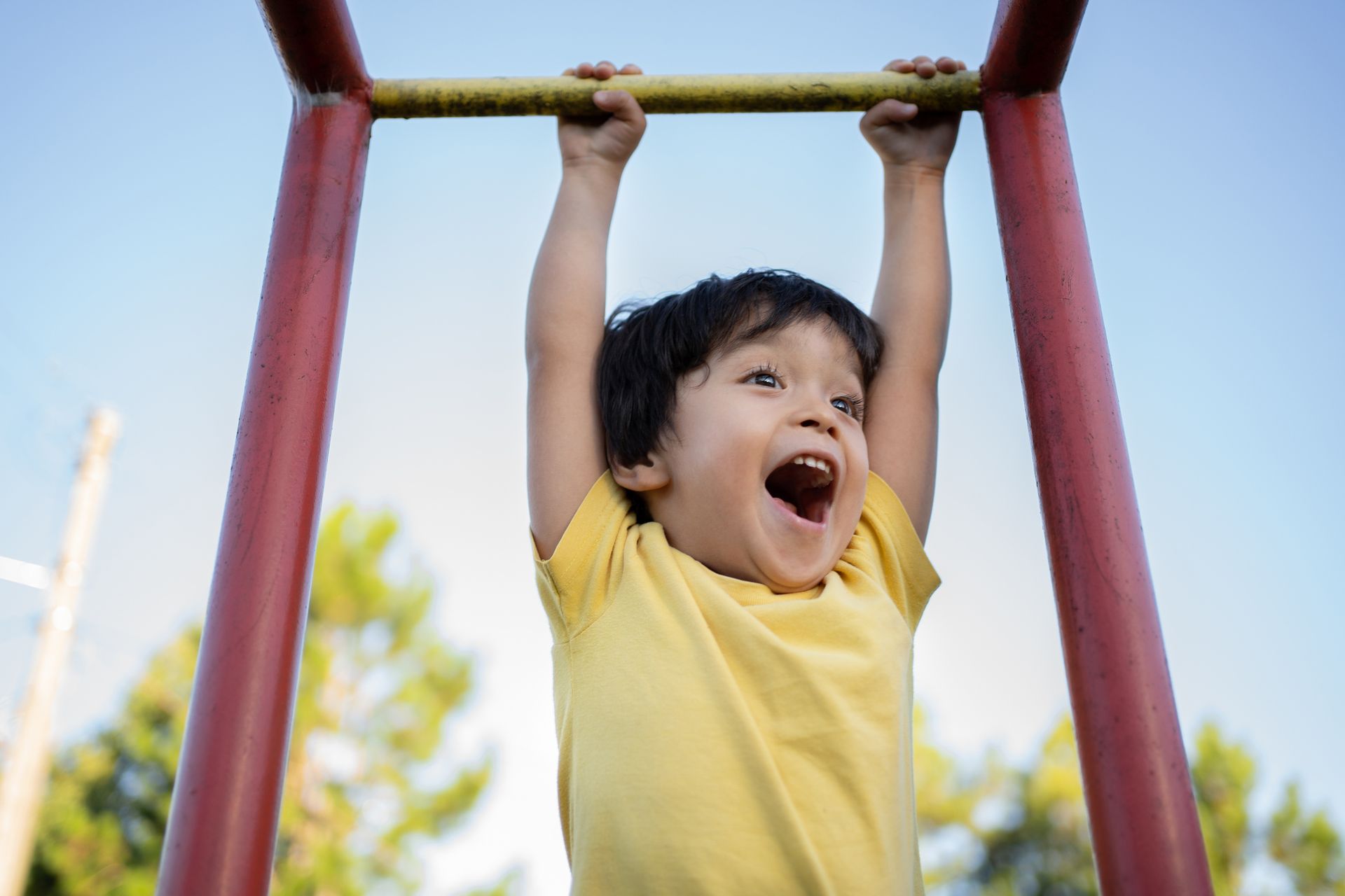 Young child hanging from monkey bars, mouth open in a joyful shout, outdoors.