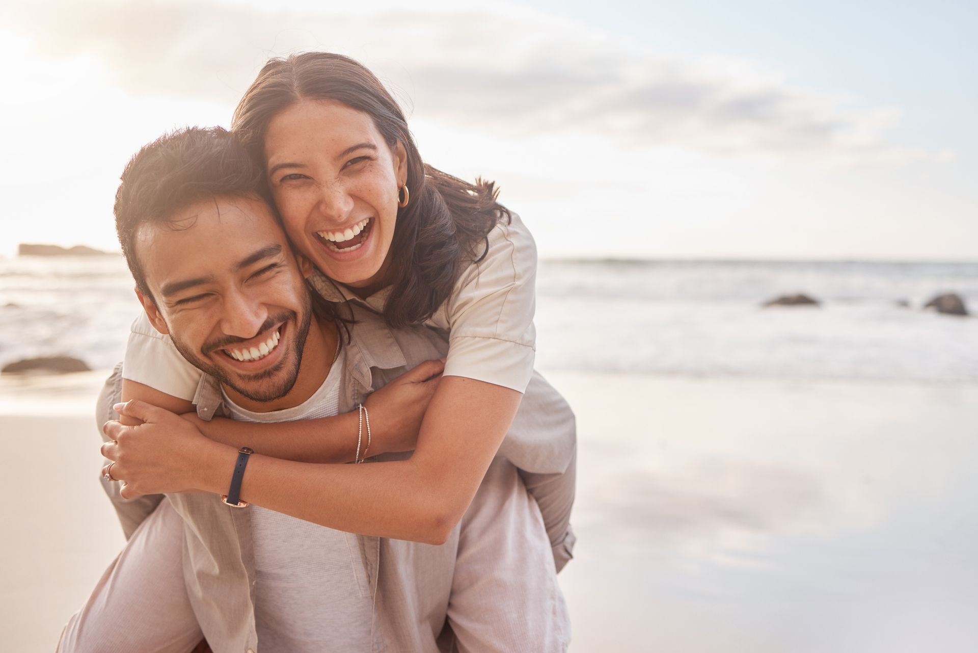 Man carrying a woman piggyback on a beach, both laughing. Golden sunlight.