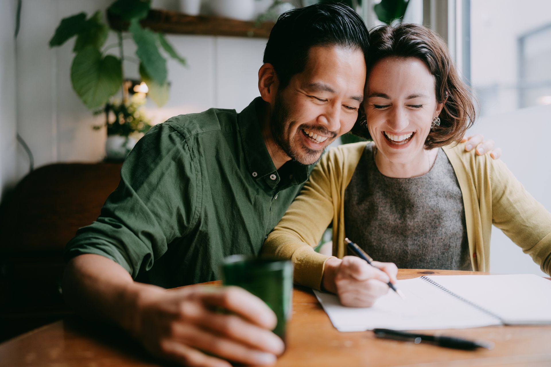 Man and woman laughing while writing at a table by a window, green and yellow clothing.