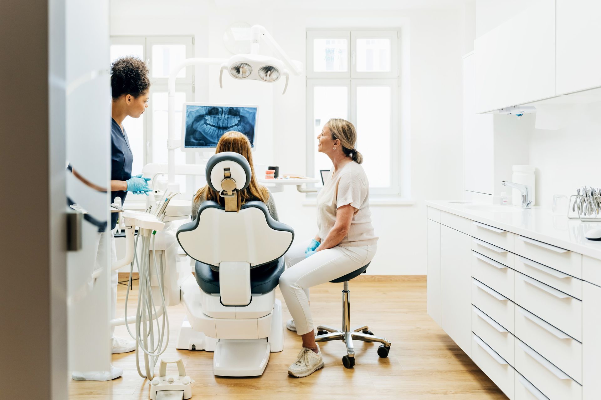 Dentist examining a patient in a dental chair. Assistant and X-rays are visible. Bright, modern office.