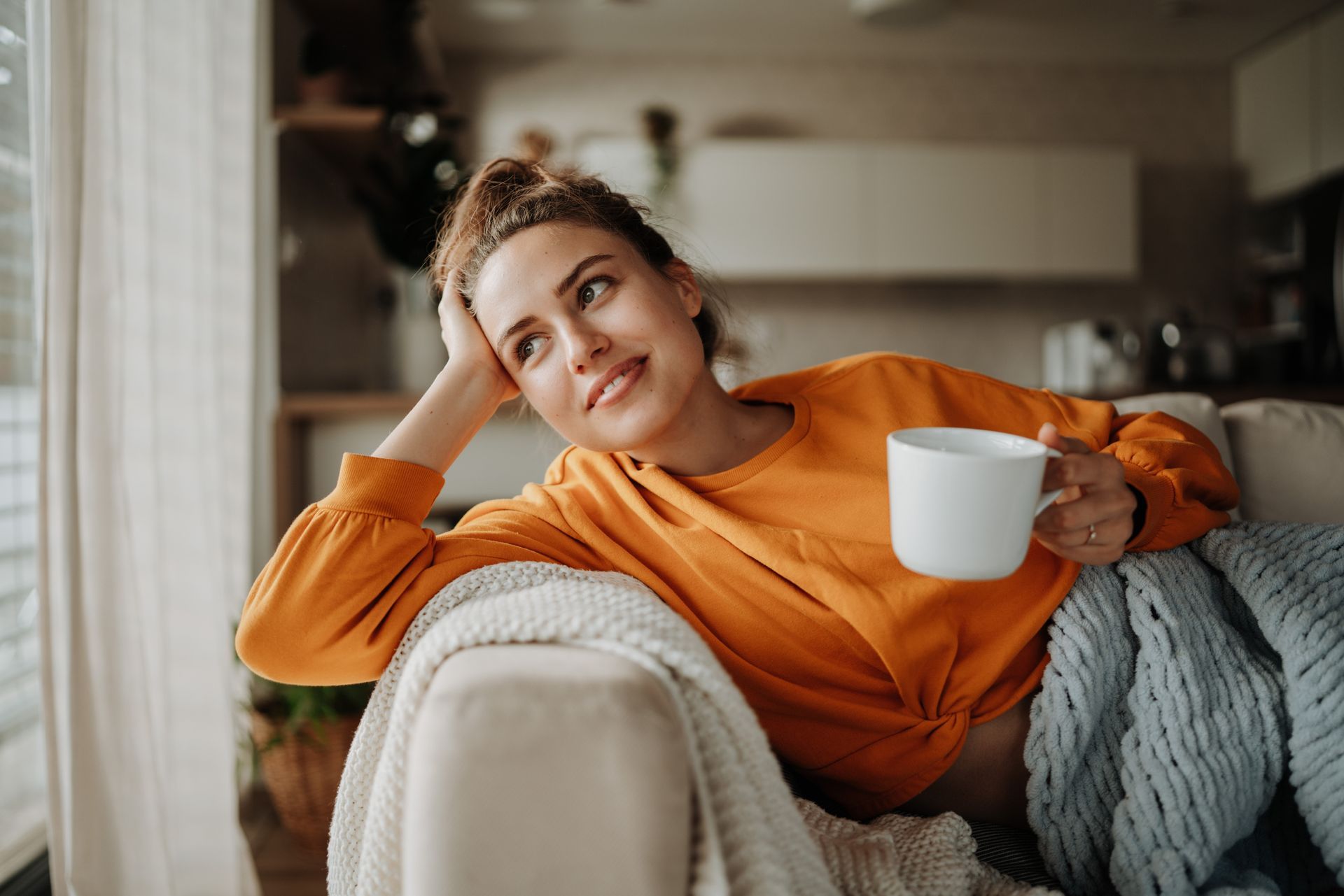 Woman on a couch smiles, holding a mug, looking out a window in a cozy living room setting.