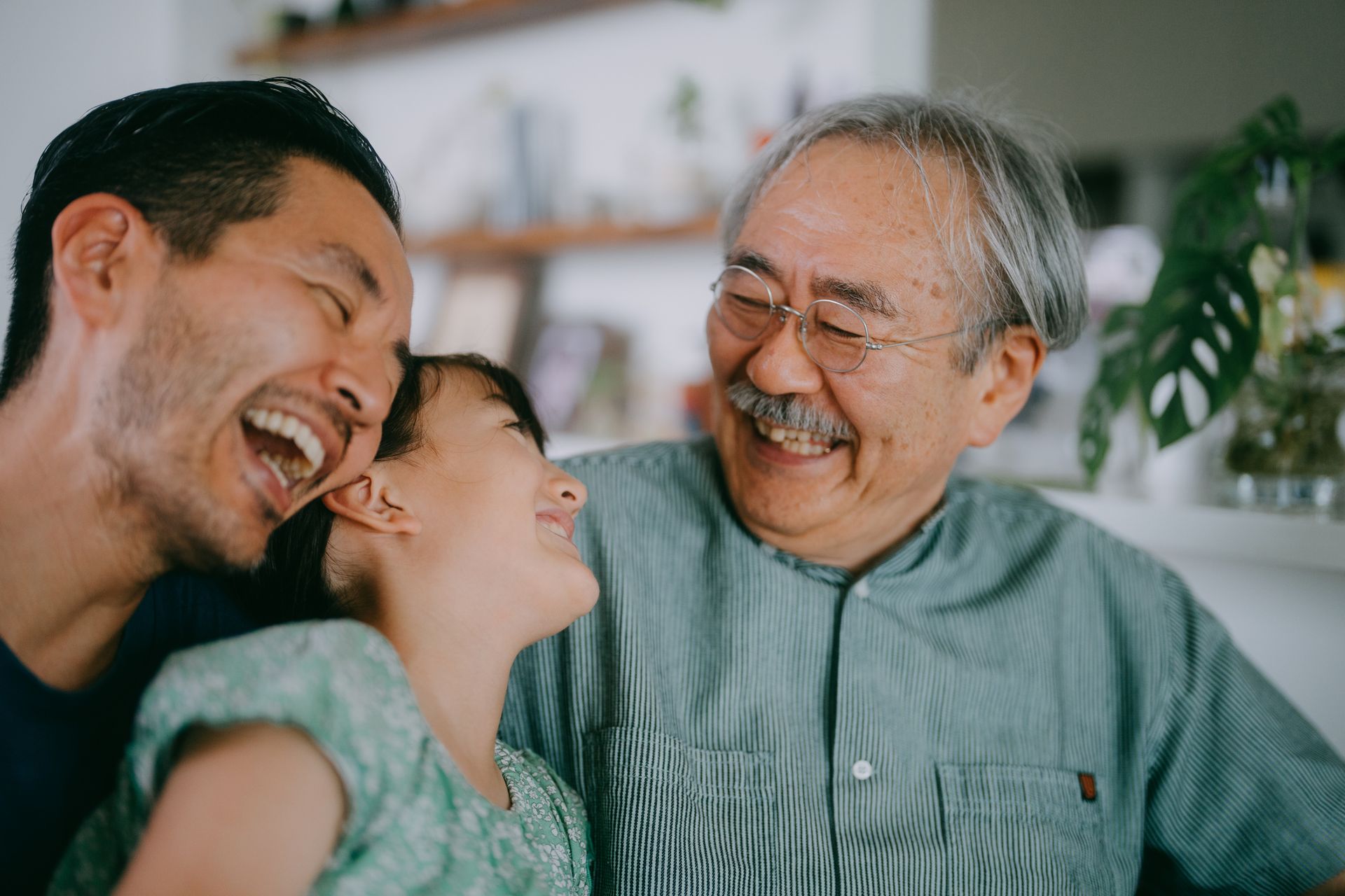 A smiling Asian family: father, daughter, and grandfather, indoors. They are laughing and looking at each other.