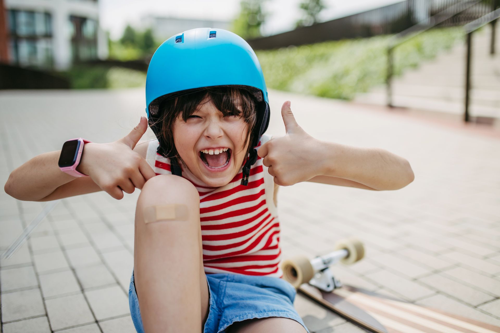 Boy in a blue helmet and striped shirt gives thumbs up, seated with a skateboard.