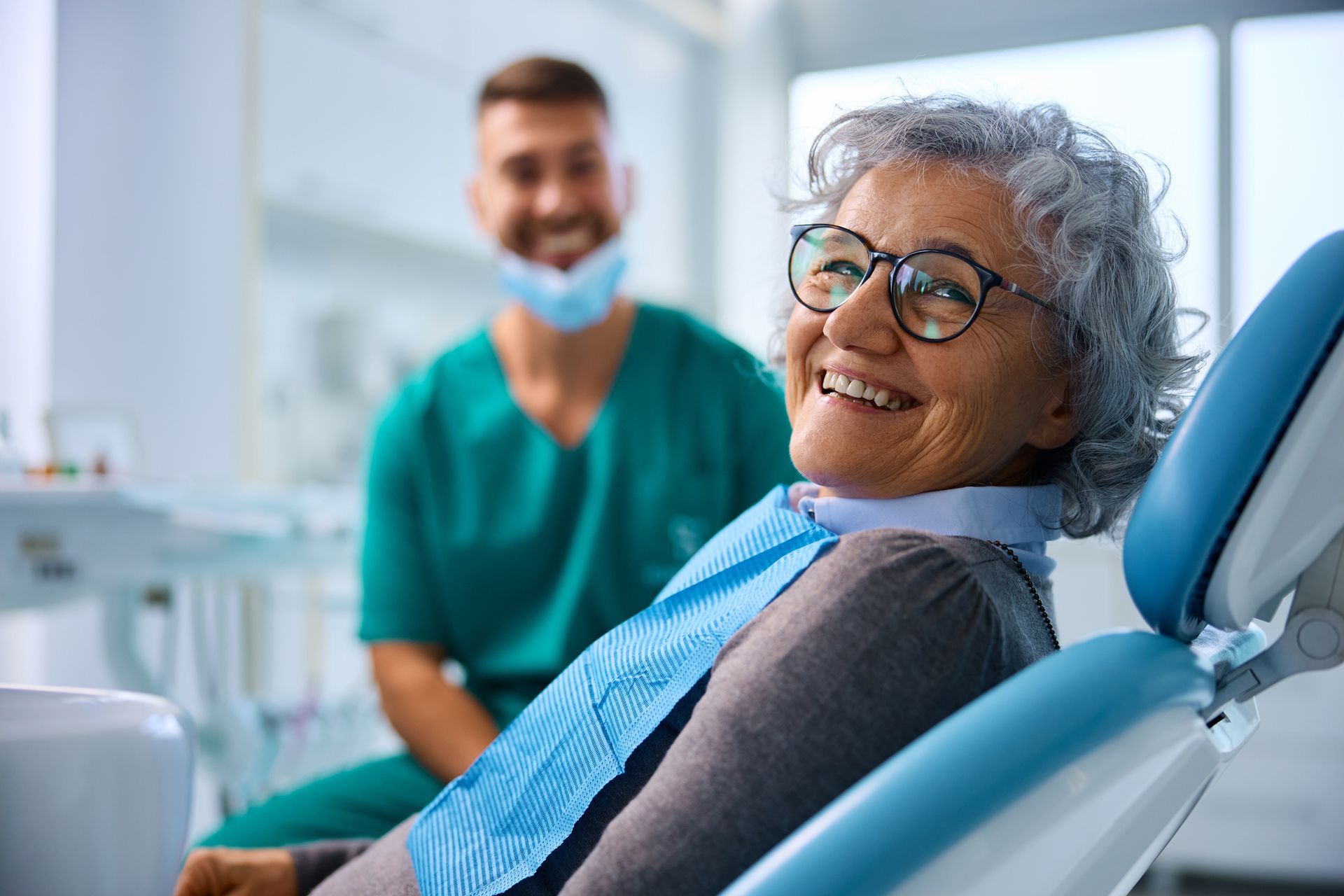 Smiling senior woman in dentist chair, dentist in scrubs in background.