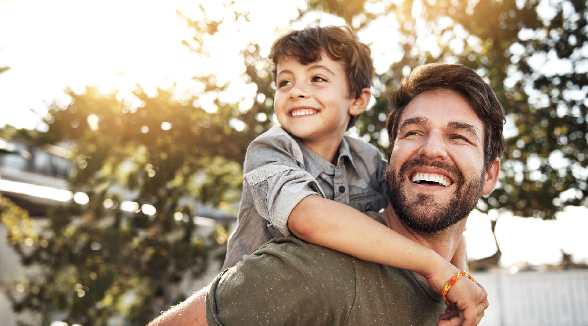 Father giving a piggyback ride to his smiling son in a sunny outdoor setting.