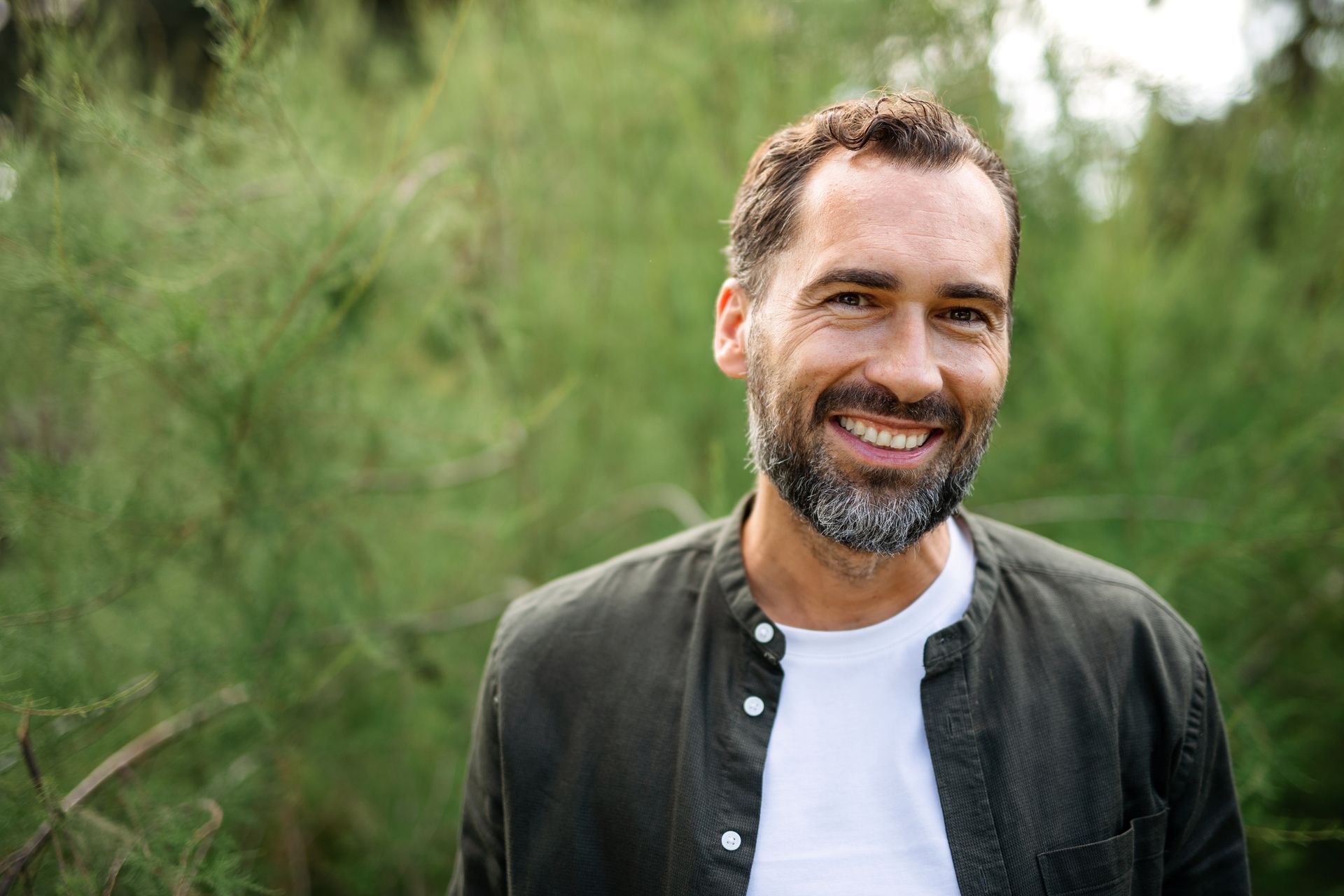 Man with brown hair and beard smiles outdoors, wearing a white shirt under a green jacket, in front of green foliage.