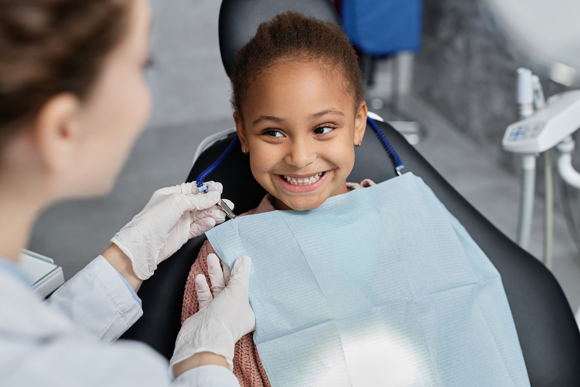 A young girl smiles at a dentist in a dental chair, wearing a protective bib.