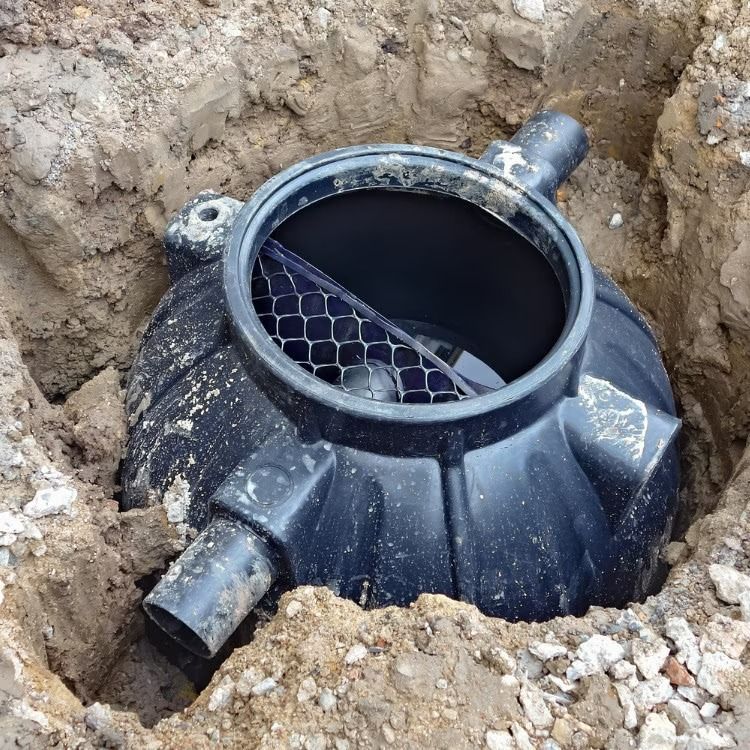 Black plastic septic tank buried in dirt, with open circular top and side pipes visible