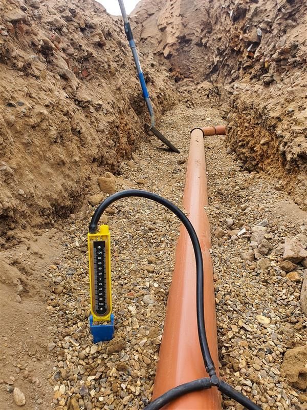 Trench with an orange pipe being installed and a yellow measuring device on a black cable