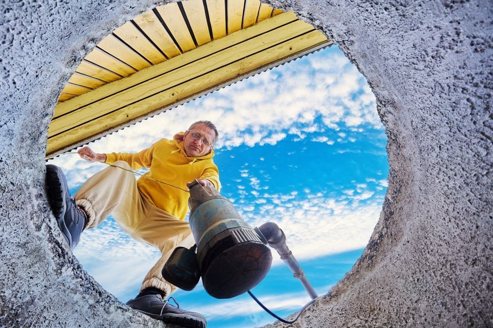 Person looking down into a circular opening, reaching with a black tool against a bright blue sky.