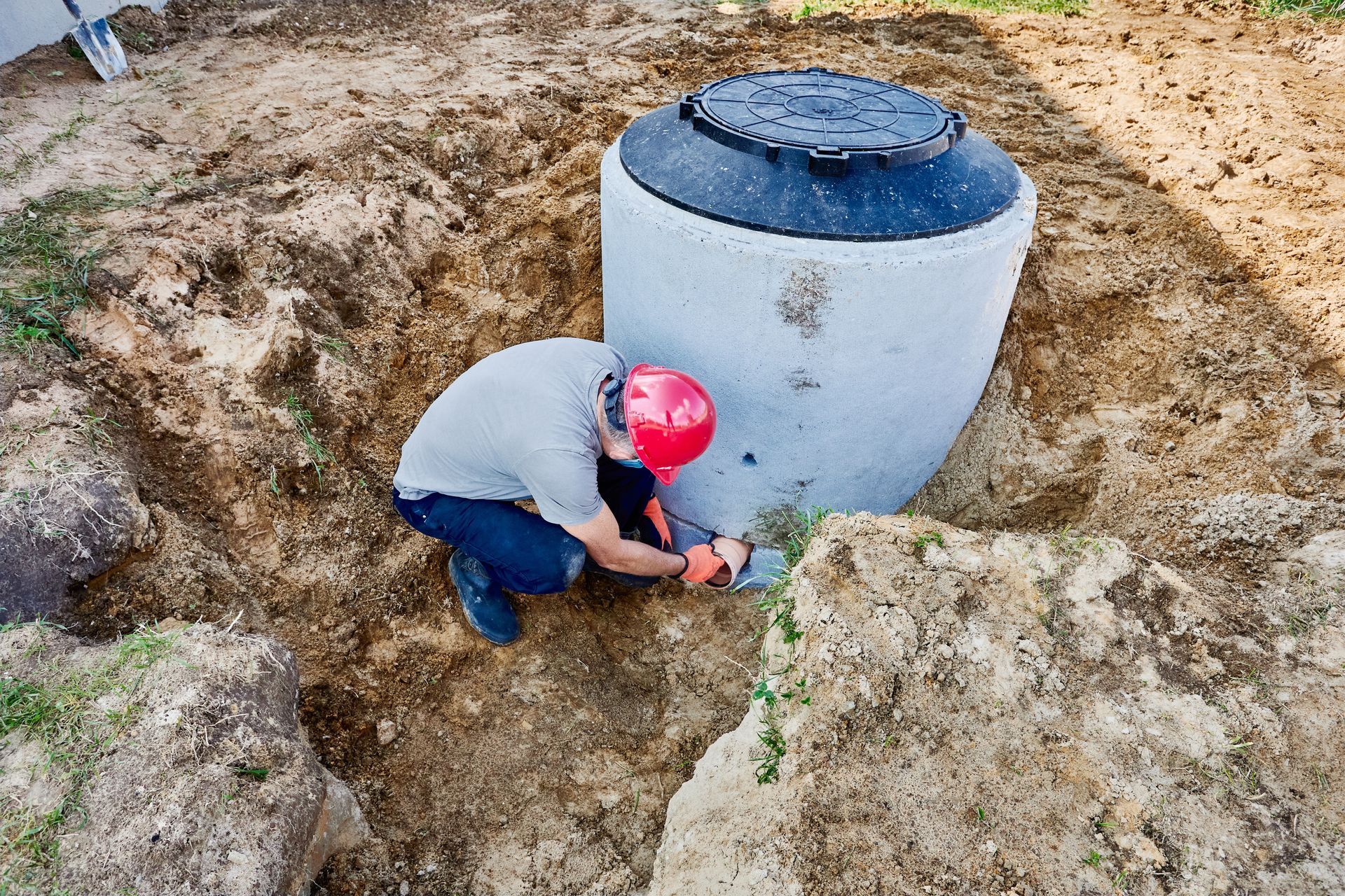 Worker in a red hard hat crouches beside a concrete tank in an excavated trench.