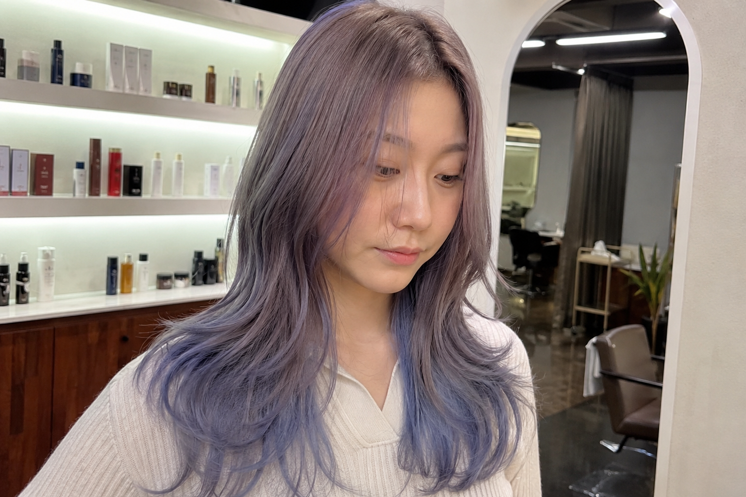 Woman with long lavender hair in a salon, looking down beside shelves of hair products at busan gwangalli haeundae