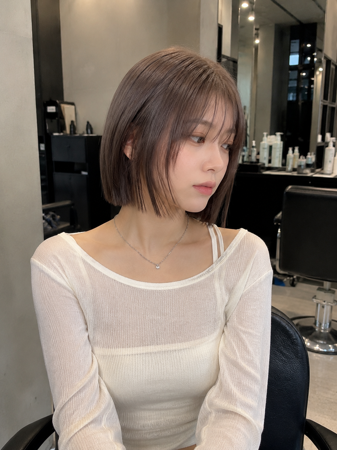 Woman with a short brown bob in a white sheer top, seated in a salon and looking to the side at busan gwangalli haeundae