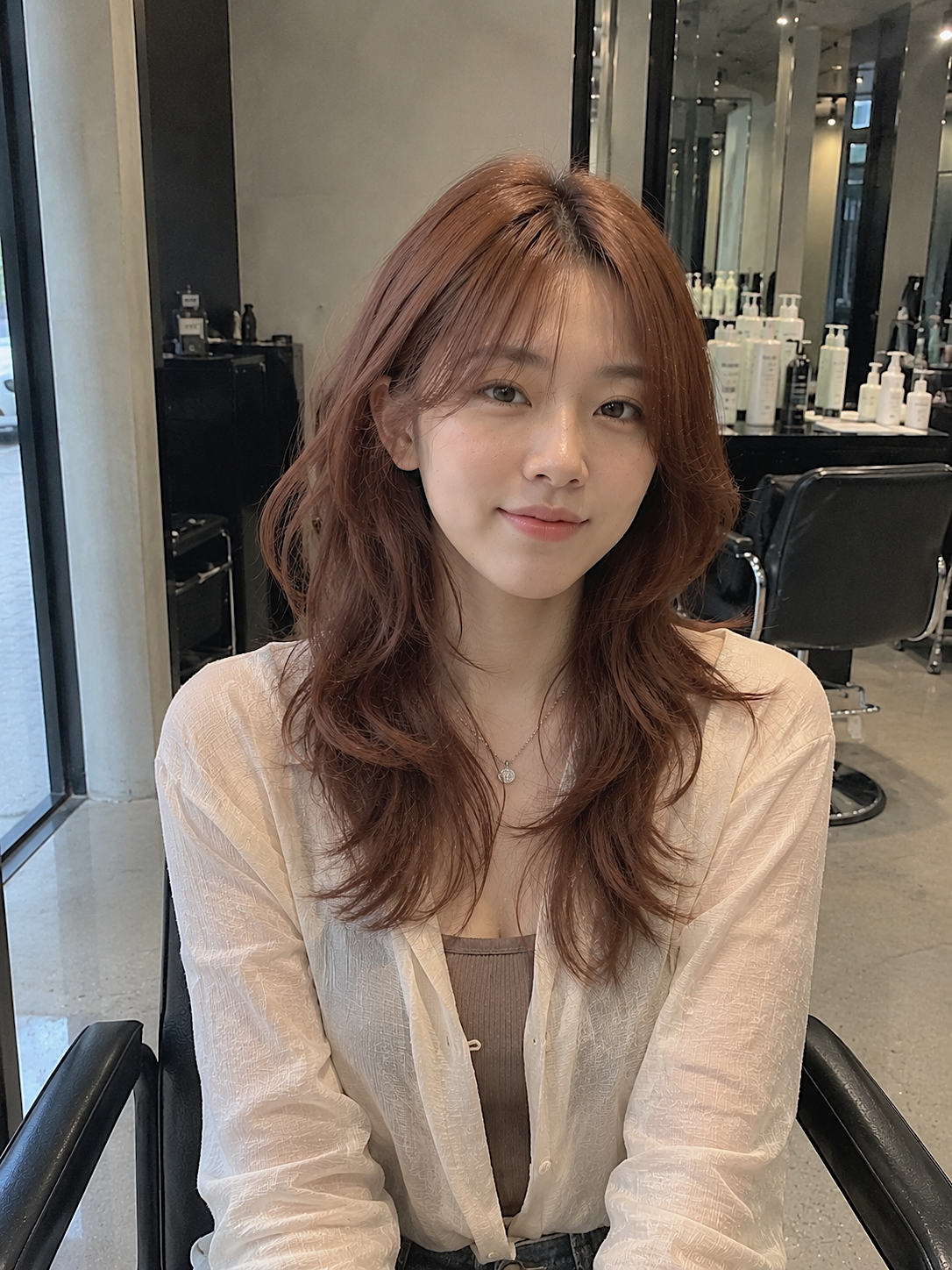 Woman with wavy auburn hair seated in a salon chair, wearing a light cardigan and smiling gently. at busan gwangalli haeundae