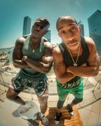 Two men lean toward the camera on a sunny city street, arms crossed, with tall buildings behind them.