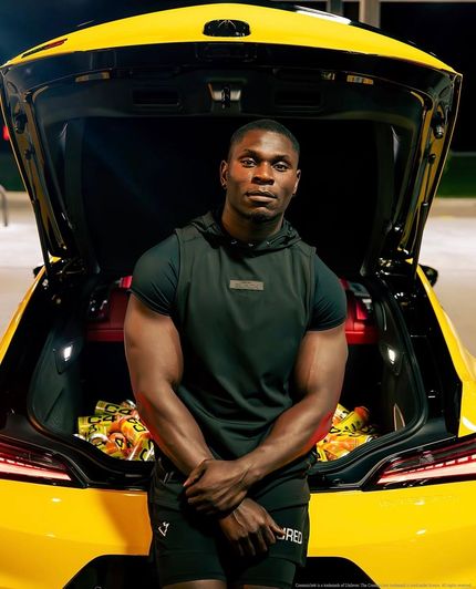 Man standing in the open trunk of a yellow sports car, wearing a dark shirt and shorts, looking at the camera