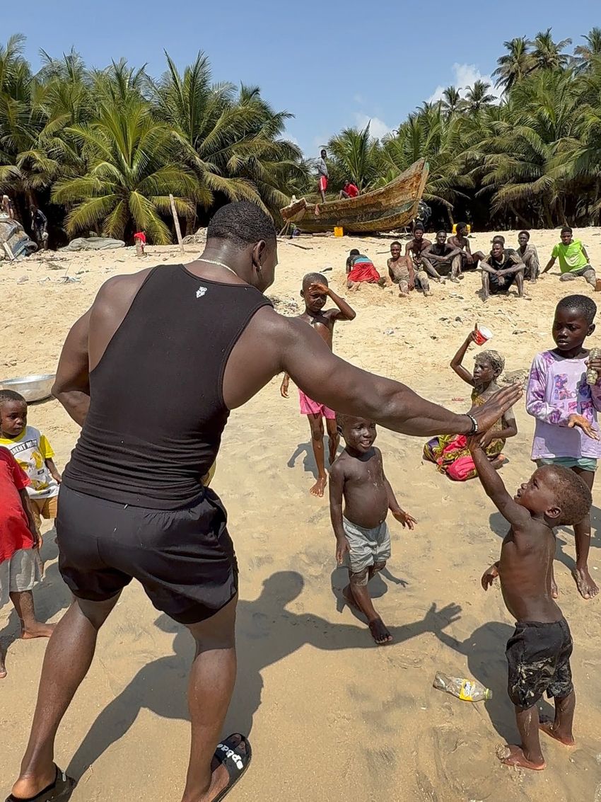 Man in black shorts dancing with children on a sandy beach, with palm trees and nets in the background