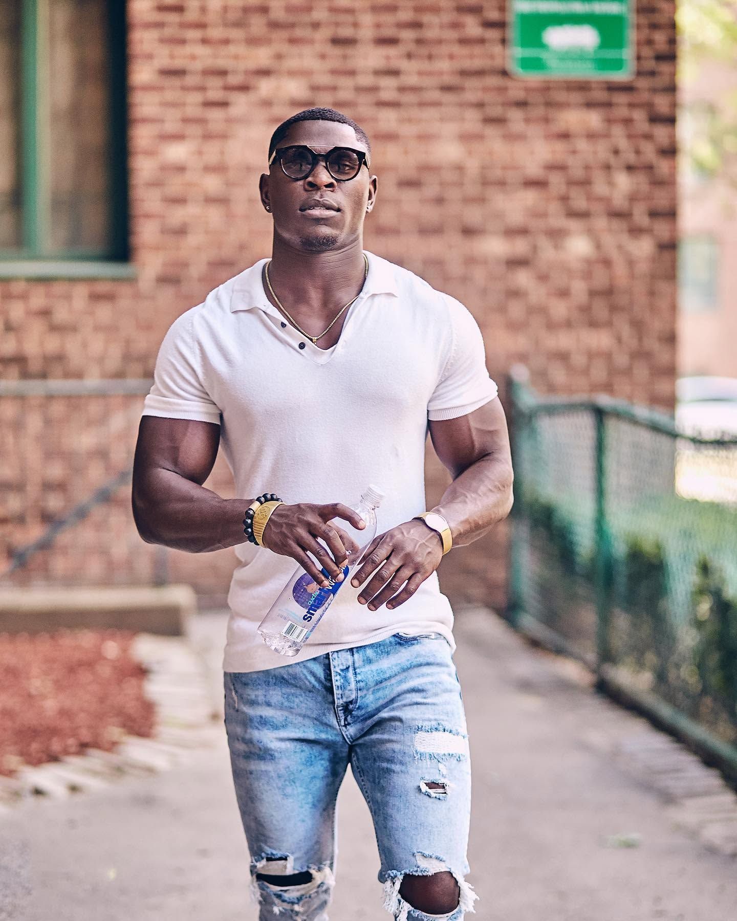 Man in sunglasses and white polo walking outdoors on a sidewalk, holding a water bottle.