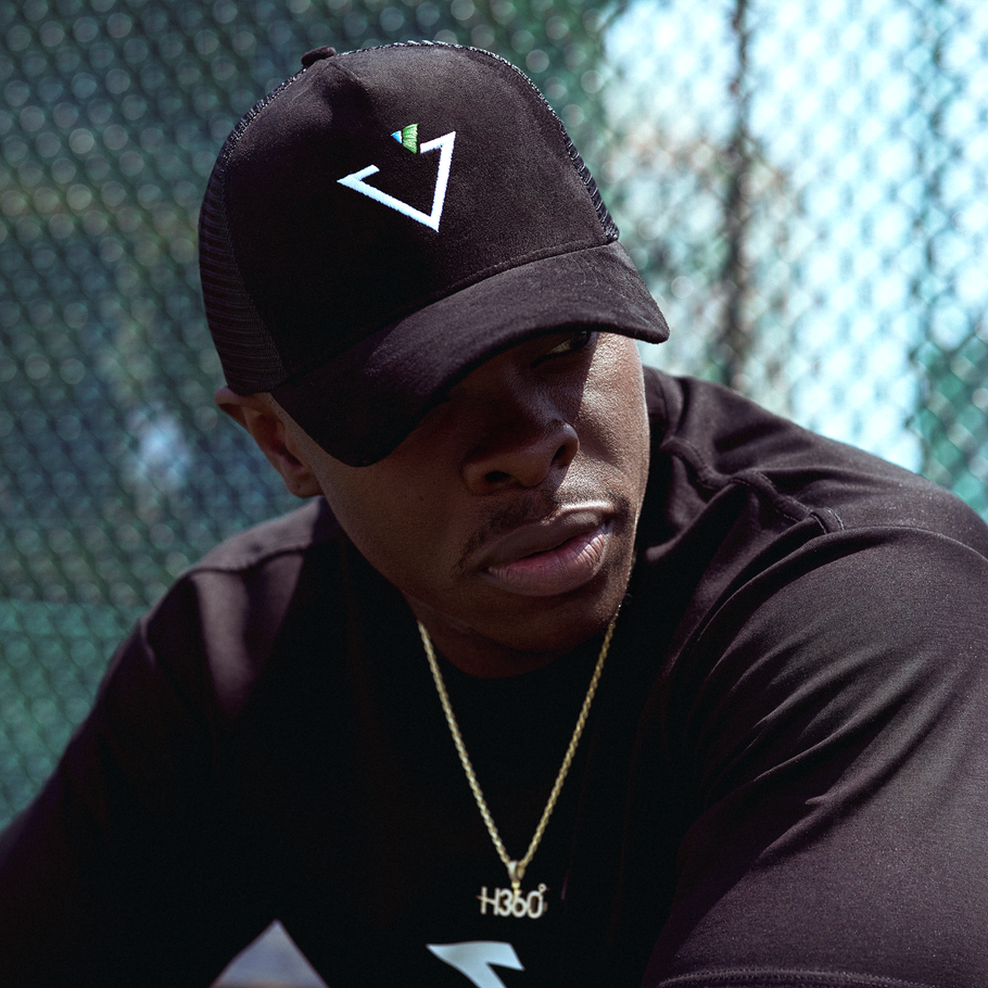 Man in black cap and shirt with gold chain, looking down outdoors by a chain-link fence.